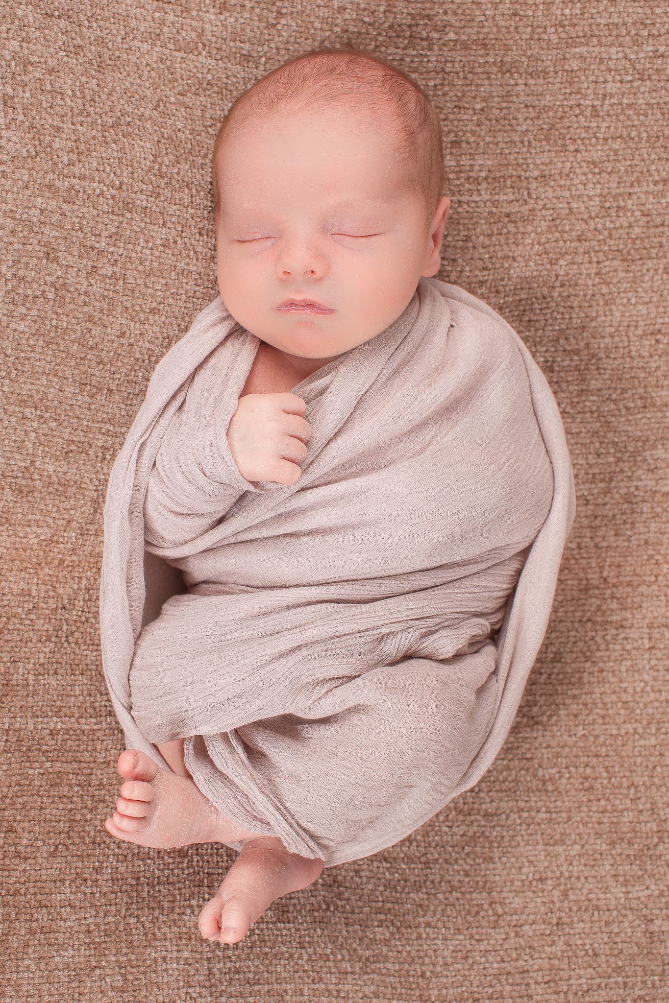 Sleeping newborn baby swaddled snugly in a soft gray wrap on a brown blanket.