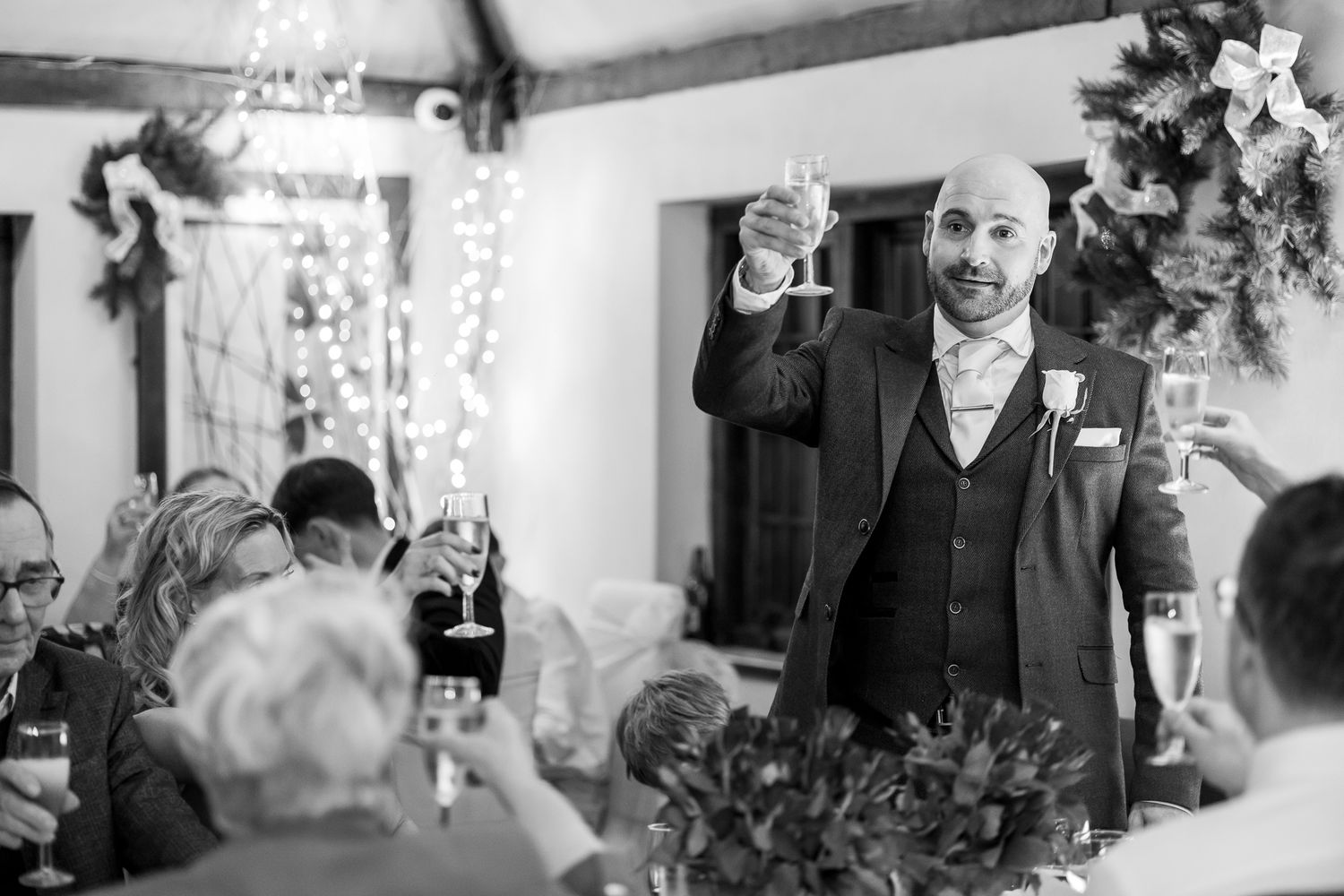 Bald groom raises champagne glass during wedding toast in black and white reception photo.