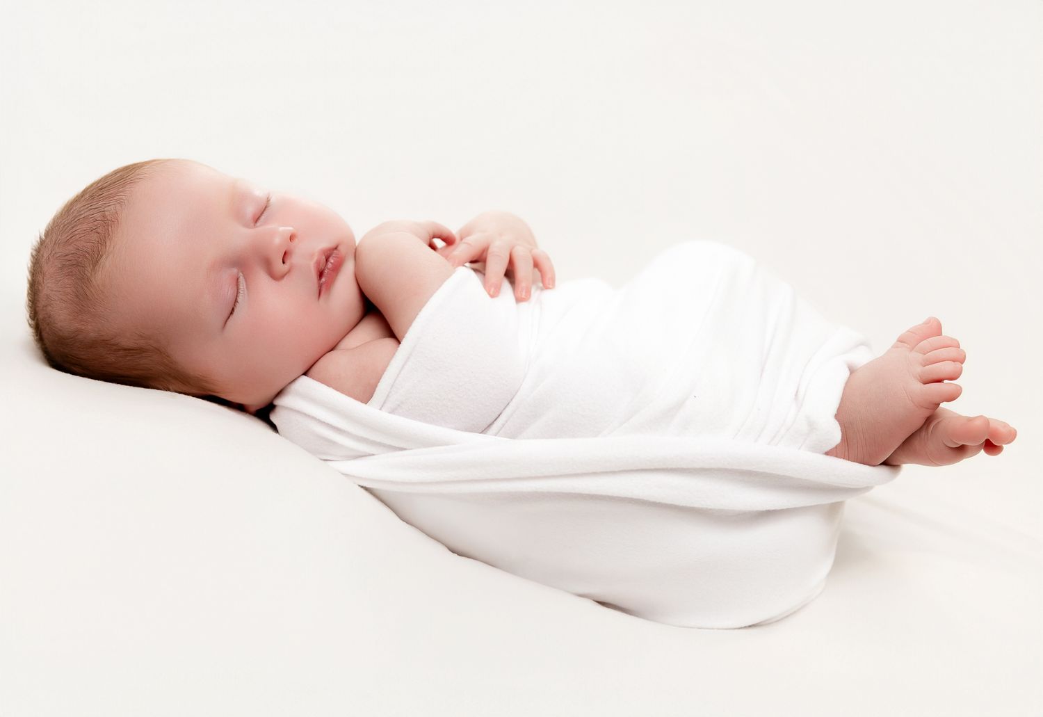 Peaceful newborn baby sleeping in a white swaddle wrap on a soft white background.