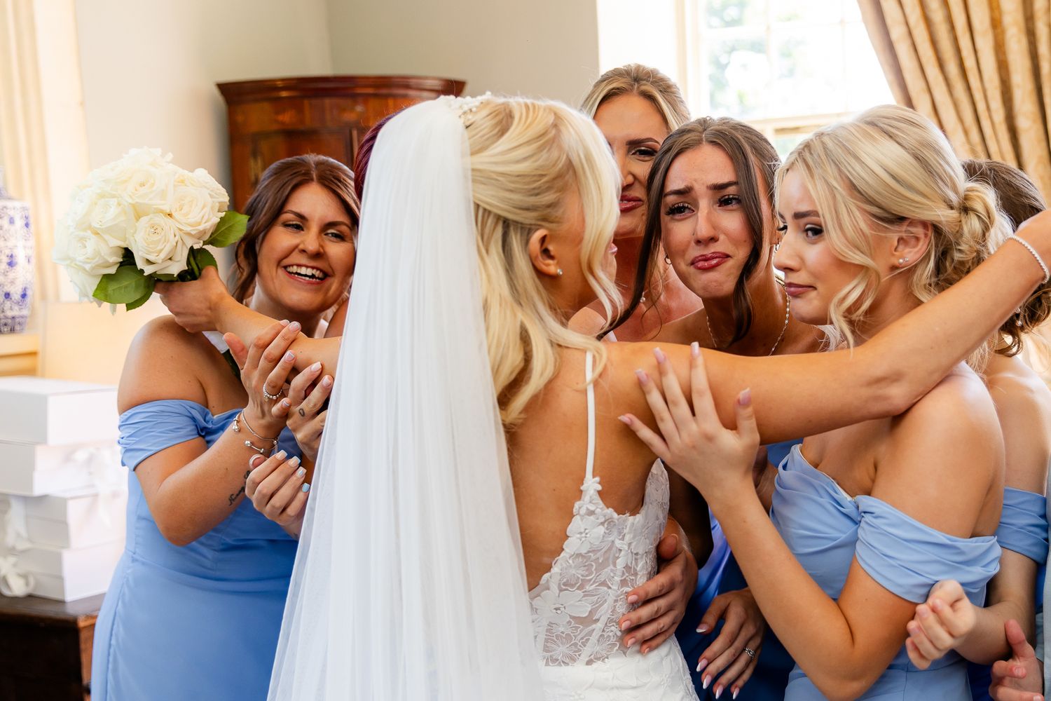 Emotional bride embraced by bridesmaids in blue dresses holding white bouquet indoors.