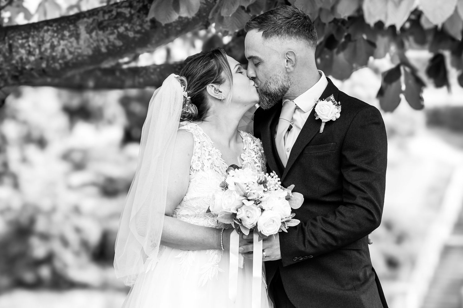 Black and white wedding photo of bride and groom sharing a kiss outdoors, bride holding a bouquet.