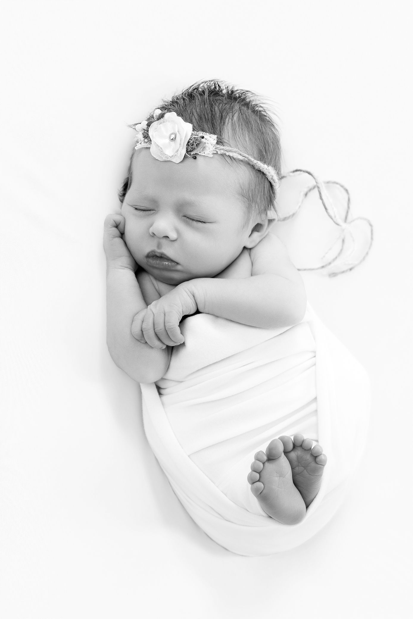 Black and white photo of a sleeping newborn baby girl swaddled in white cloth, wearing a delicate floral headband.