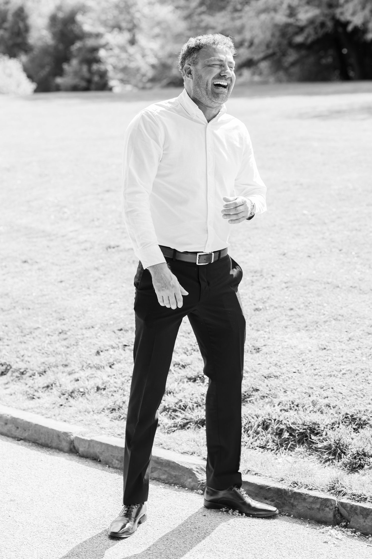 Black and white photo of a laughing man in white shirt and dress pants standing outdoors on a sunny day.