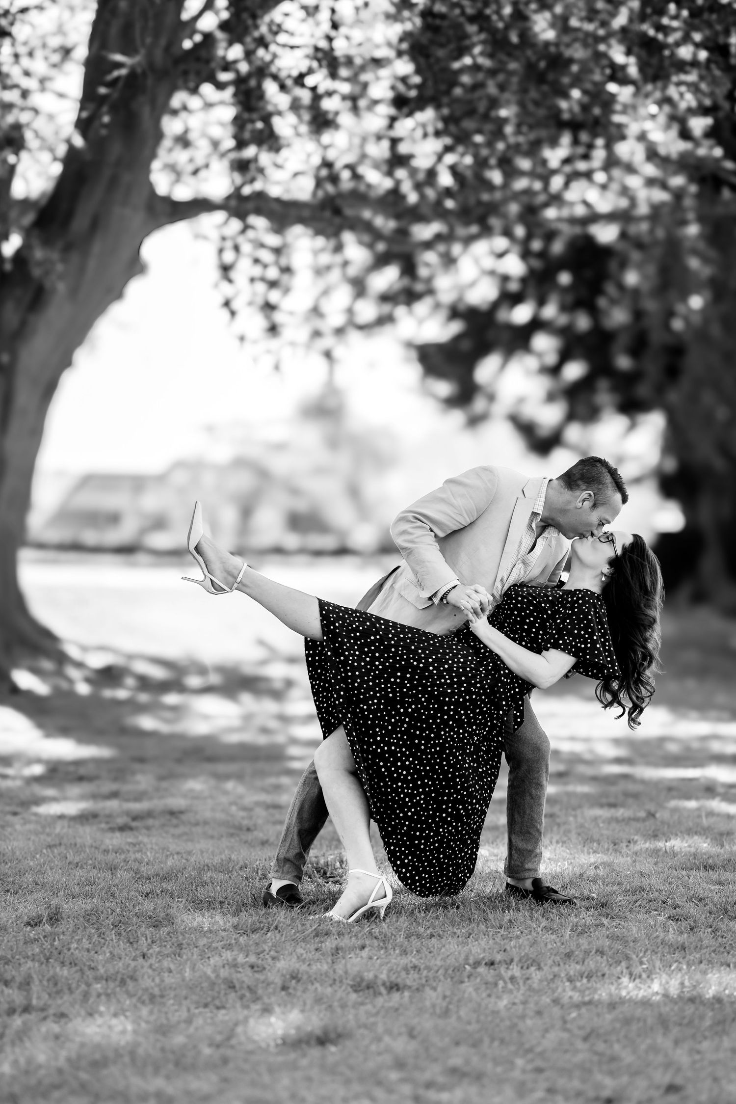 Black and white photo of a couple sharing a romantic dip kiss under a large tree on a sunny day.