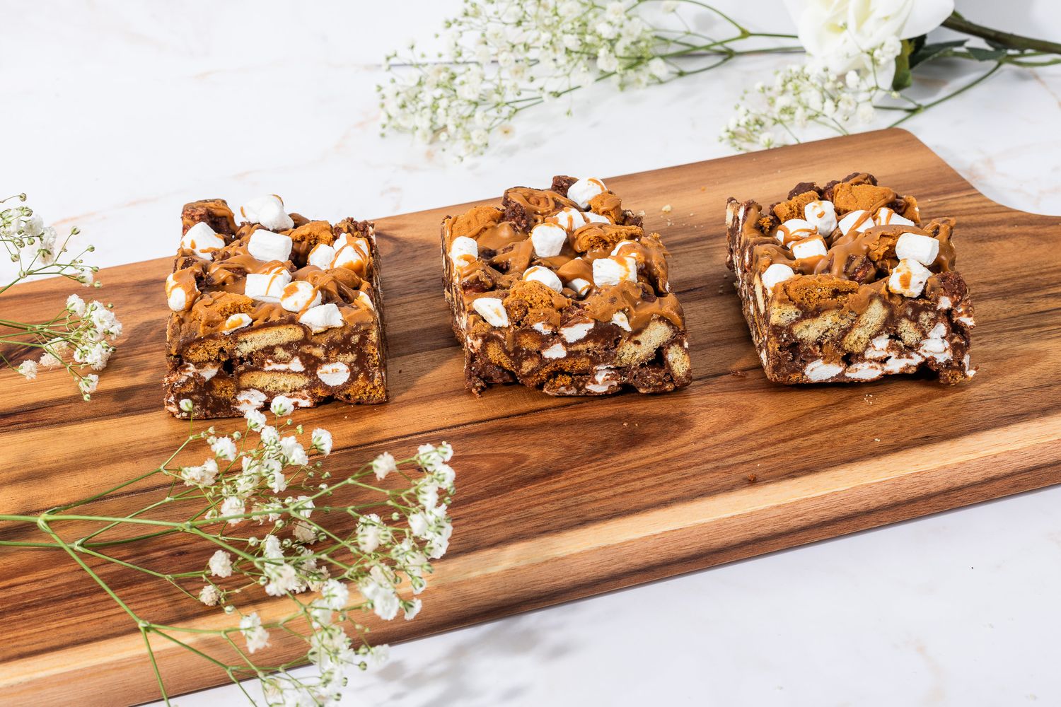 Three chocolate marshmallow brownie bars on a wooden cutting board with white flowers.