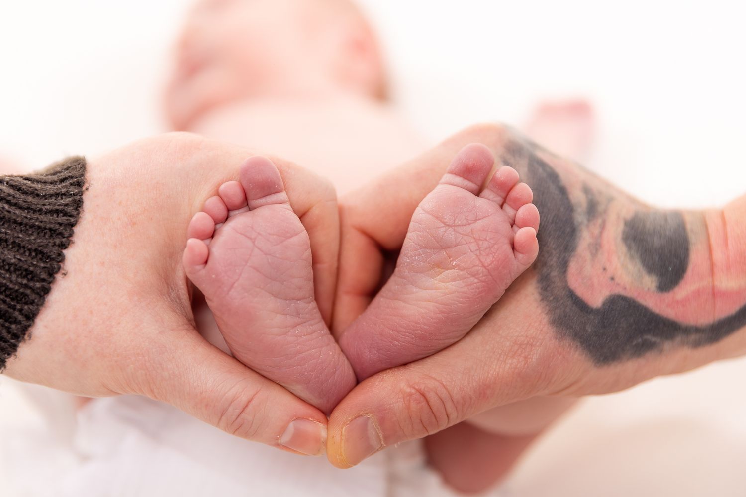 Tattooed parents hands forming a heart shape around their newborn babys tiny pink feet.