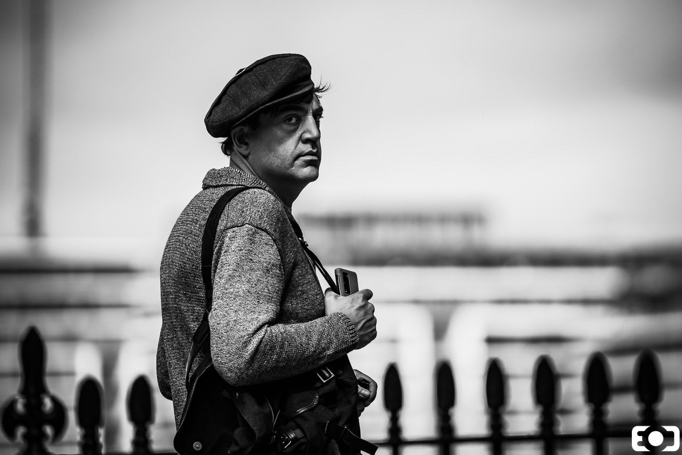 Woman wearing beret and carrying bag walking along iron fence, black and white street portrait photography.