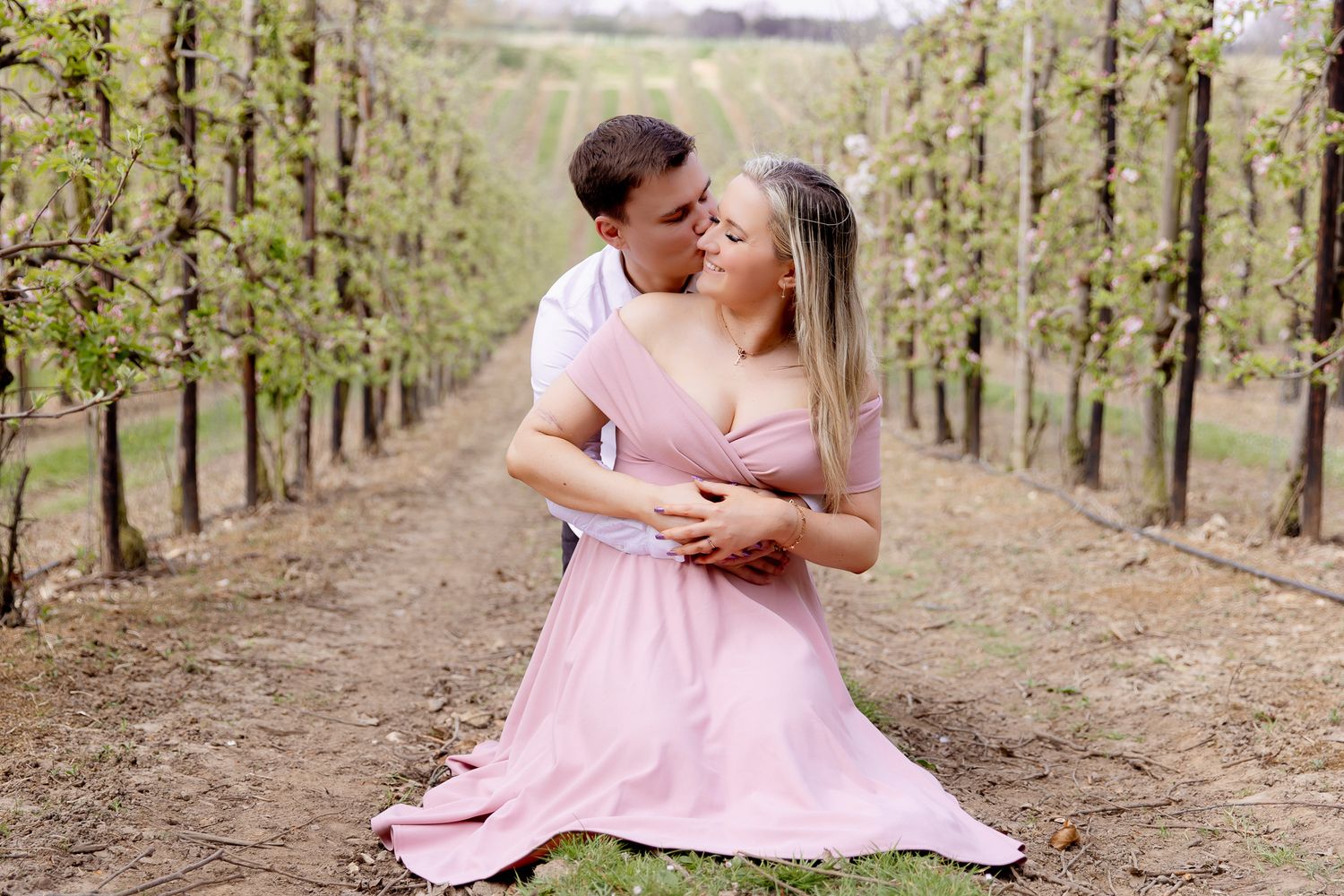 Couple sharing a tender kiss on a path between rows of trees in a vineyard.