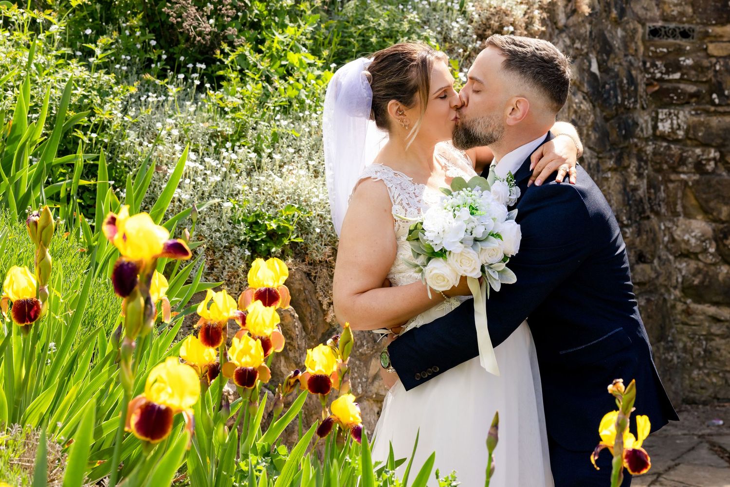 Bride and groom kissing surrounded by colorful yellow and red iris flowers in a lush garden on their wedding day.