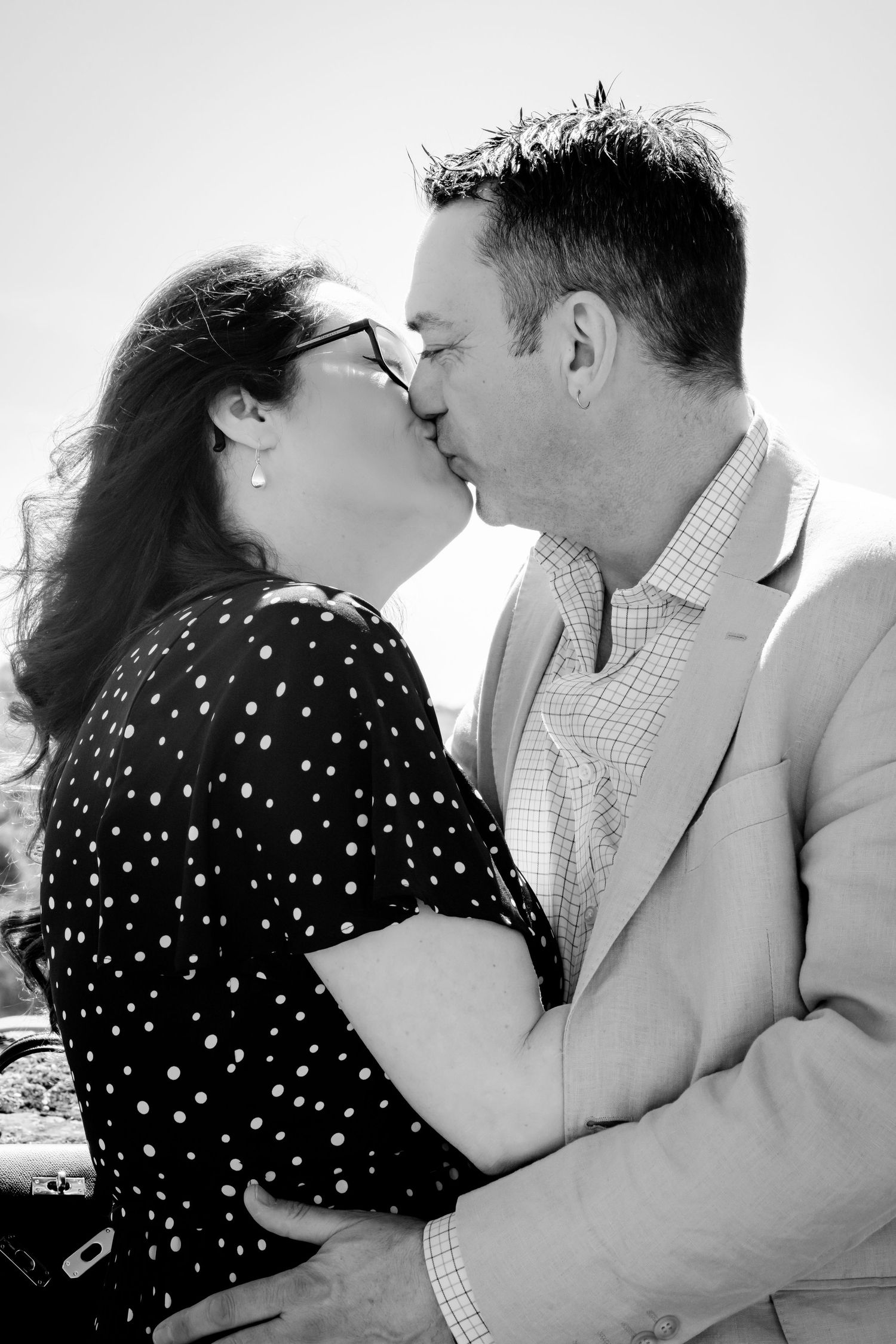 Black and white photo of a couple sharing a romantic kiss outdoors on a sunny day.