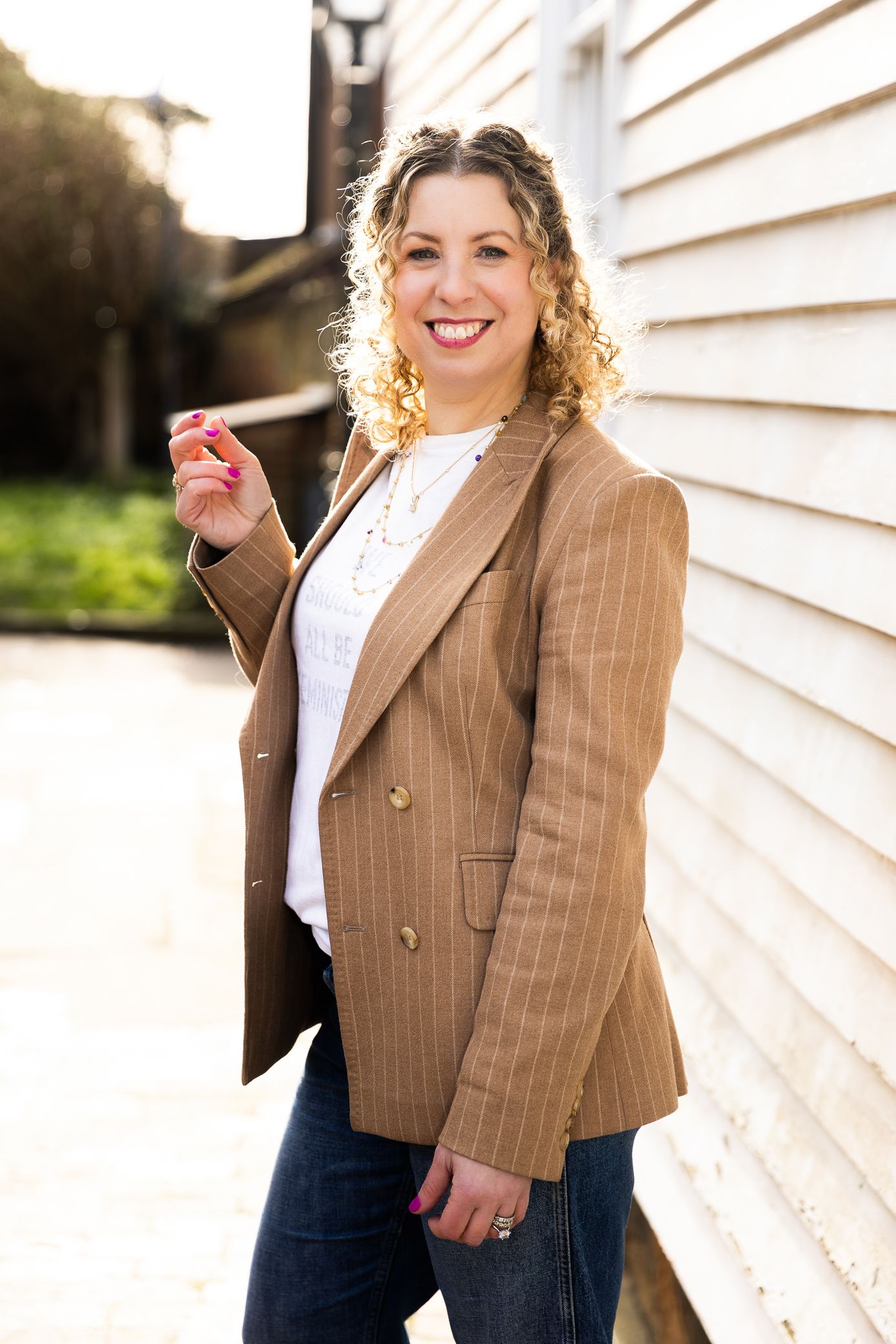 Smiling curly blonde woman in a tan blazer posing outdoors against a white wall.