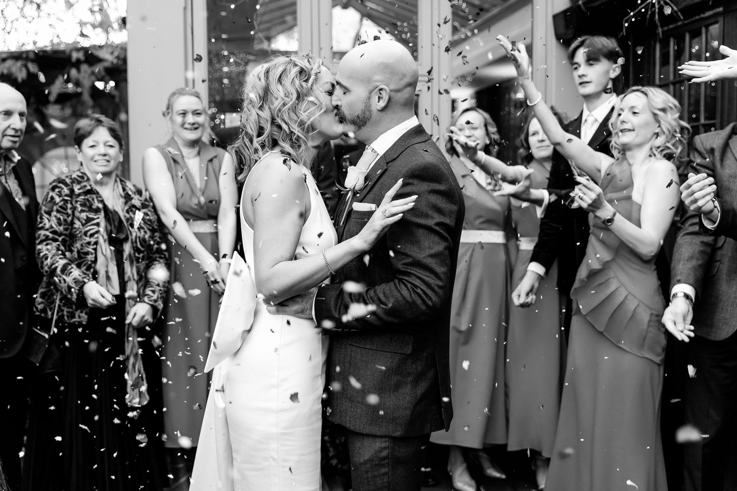 Black and white wedding photo of bride and groom sharing a kiss surrounded by guests throwing confetti.