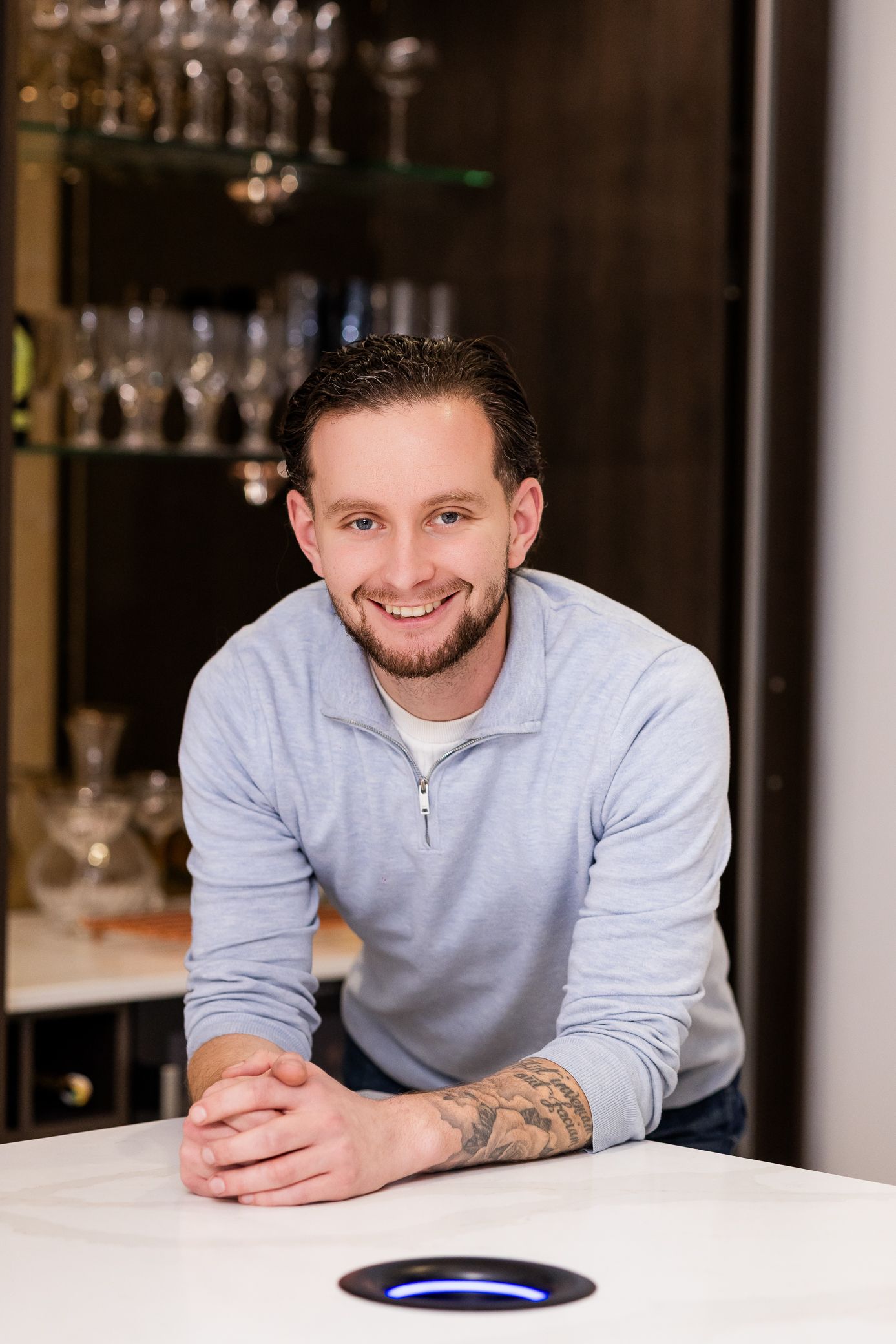 Smiling man with tattoos leaning on a white counter in a modern bar setting.