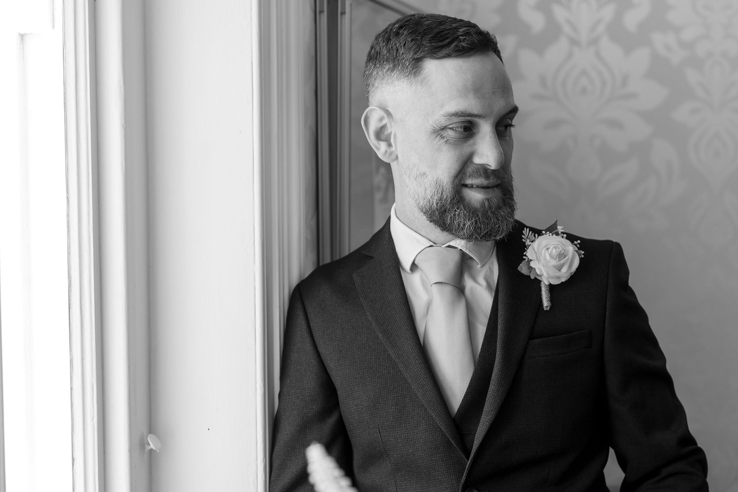 Groom in dark suit with boutonniere and tie smiling softly by window in black and white wedding photo.