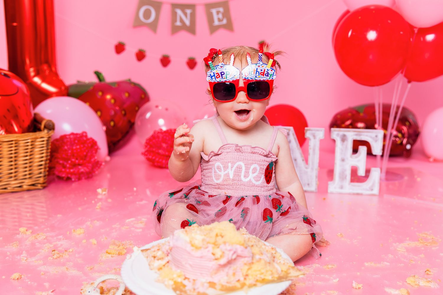 Baby girl in red sunglasses celebrating first birthday cake smash on pink background with ONE decorations.