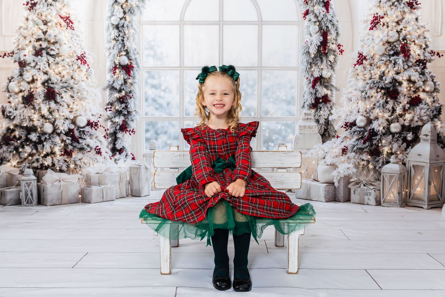 A young girl in a red plaid Christmas dress sits smiling before decorated holiday trees and gifts.