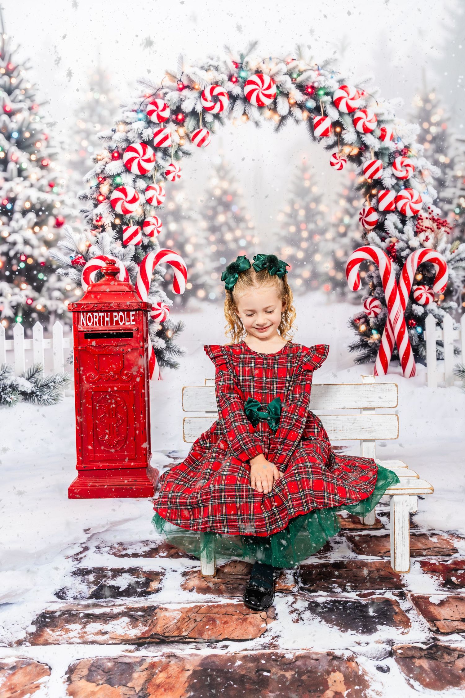 A young girl in a red plaid dress sits on a white bench near a North Pole mailbox in a snowy Christmas scene.