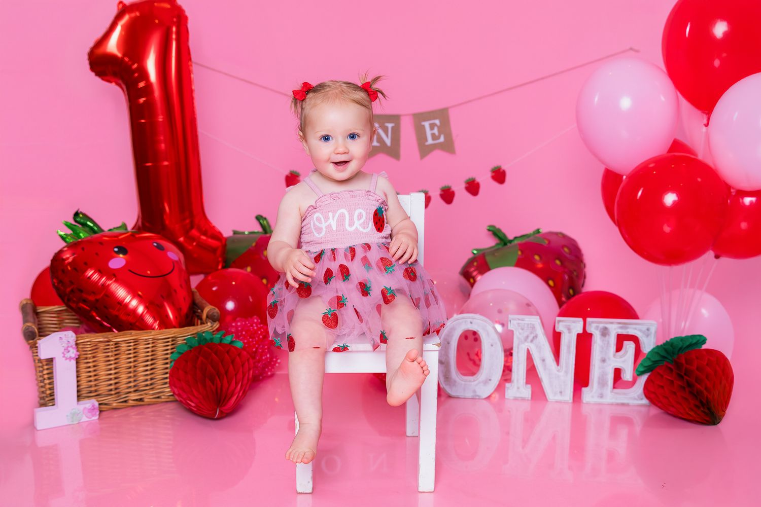 Baby girl in strawberry dress sits on white chair with red balloons and ONE decorations for first birthday cake smash.