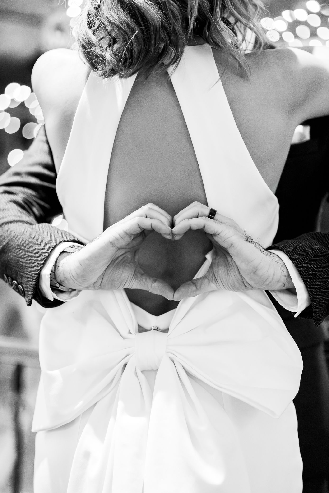 Groom makes heart shape with hands on brides open-back white wedding gown with bow.