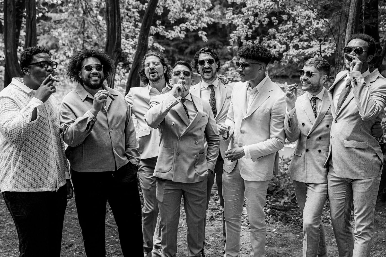 Black and white photo of a groom and groomsmen laughing together in suits among the trees.