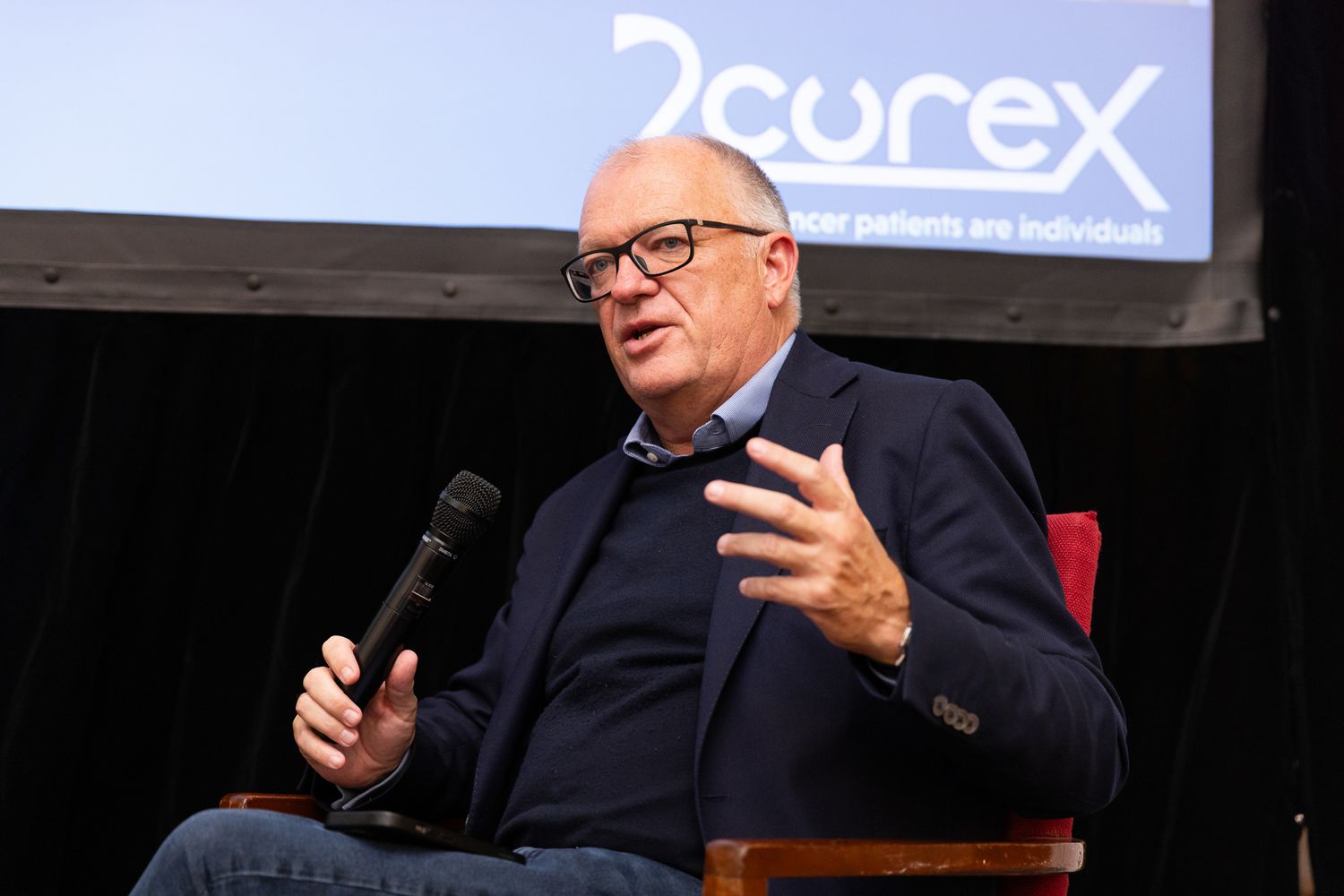 Man wearing glasses speaking passionately at a 2Curex medical conference panel discussion.