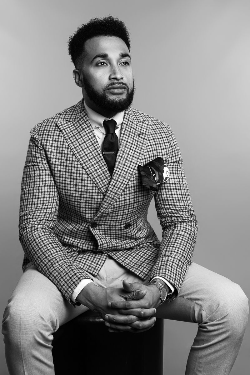 Black and white portrait of a bearded man in a plaid suit sitting confidently on a stool.