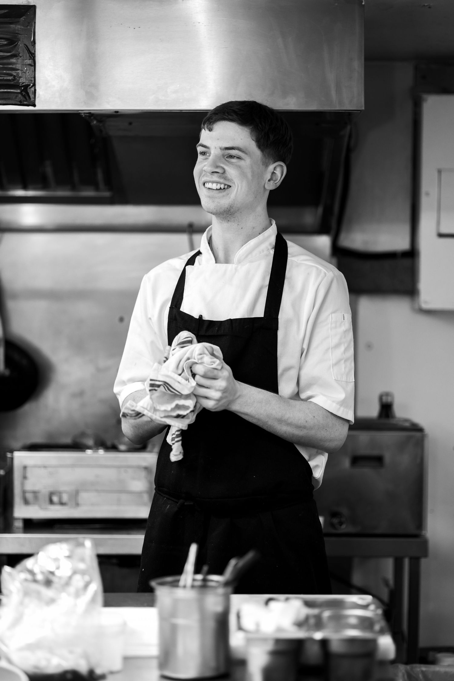 Smiling young chef in white coat and black apron drying hands with towel in professional kitchen, black and white photo.