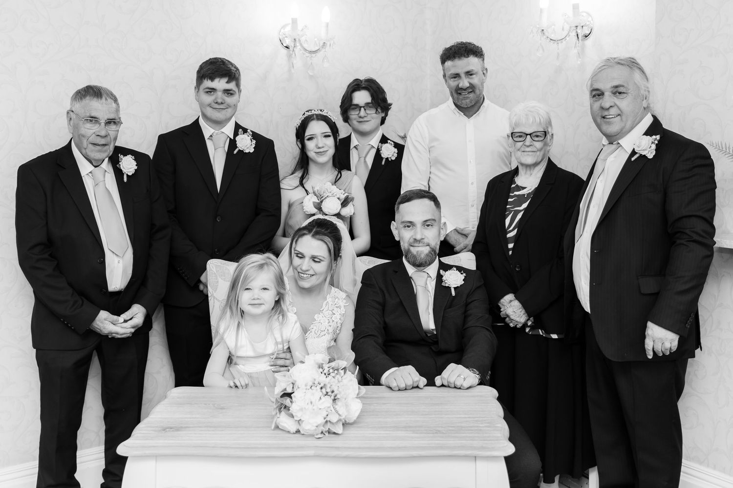 Black and white wedding photo of couple signing register surrounded by smiling family members in formal attire.