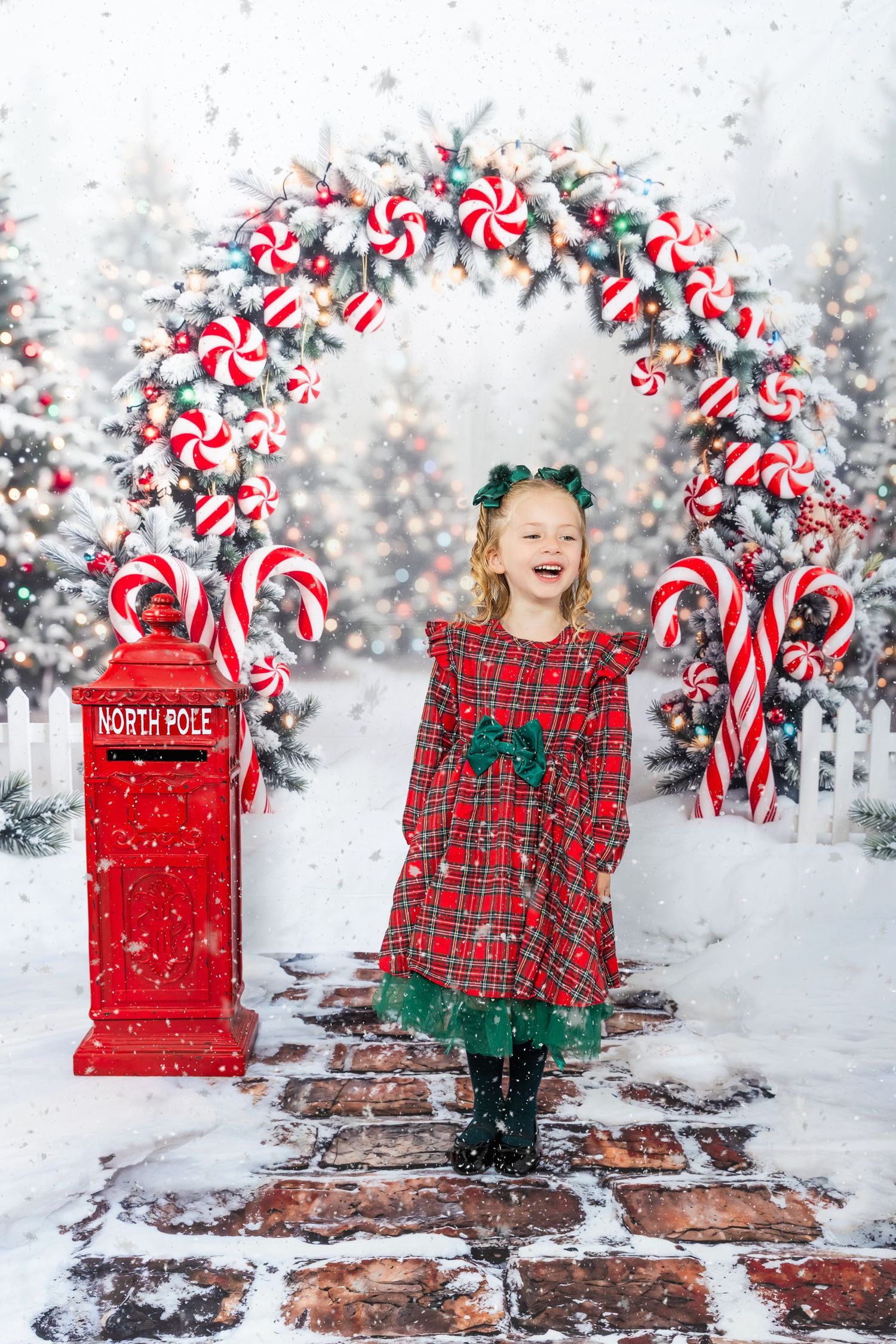 Girl in red plaid dress laughing by North Pole mailbox under candy cane arch with snowy Christmas trees.
