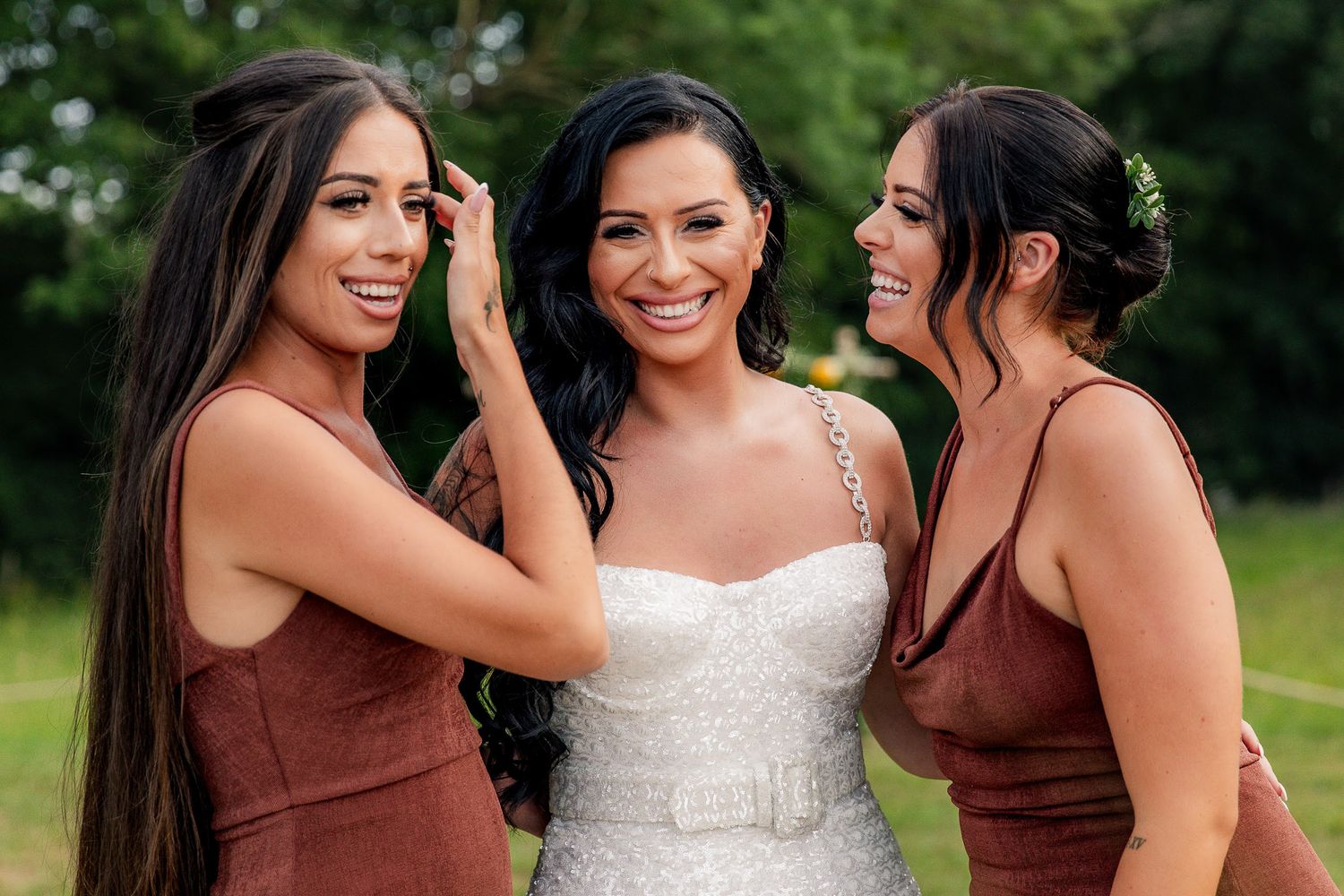 Bride in white gown flanked by two bridesmaids in dusty rose dresses outdoors.