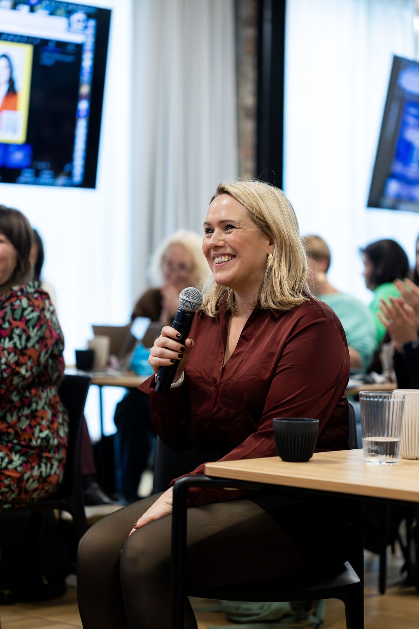 Smiling blonde woman holds microphone while seated at an indoor professional panel event.