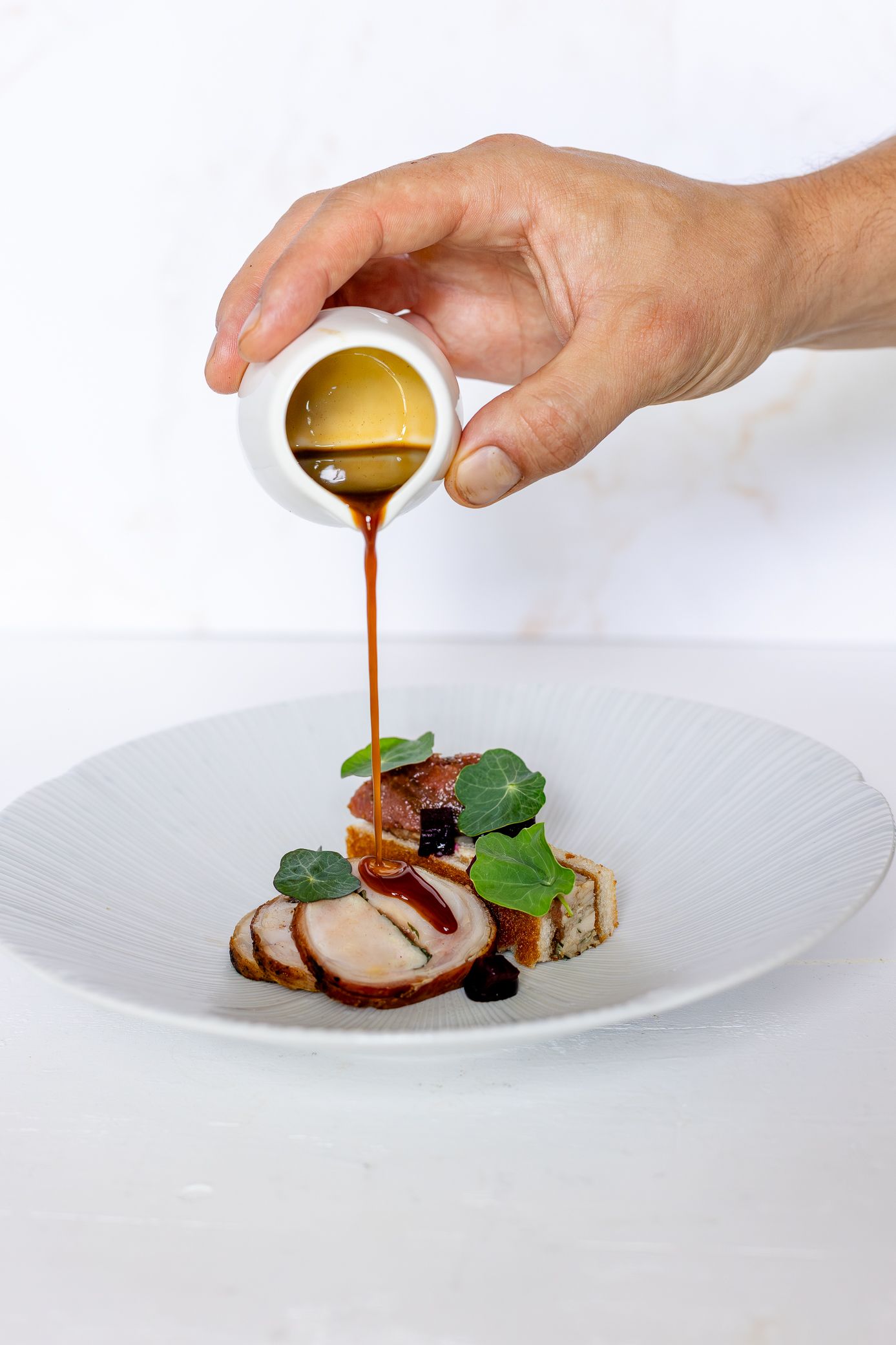 Chef pouring rich brown sauce over elegant plated meat dish with fresh herbs on white plate.