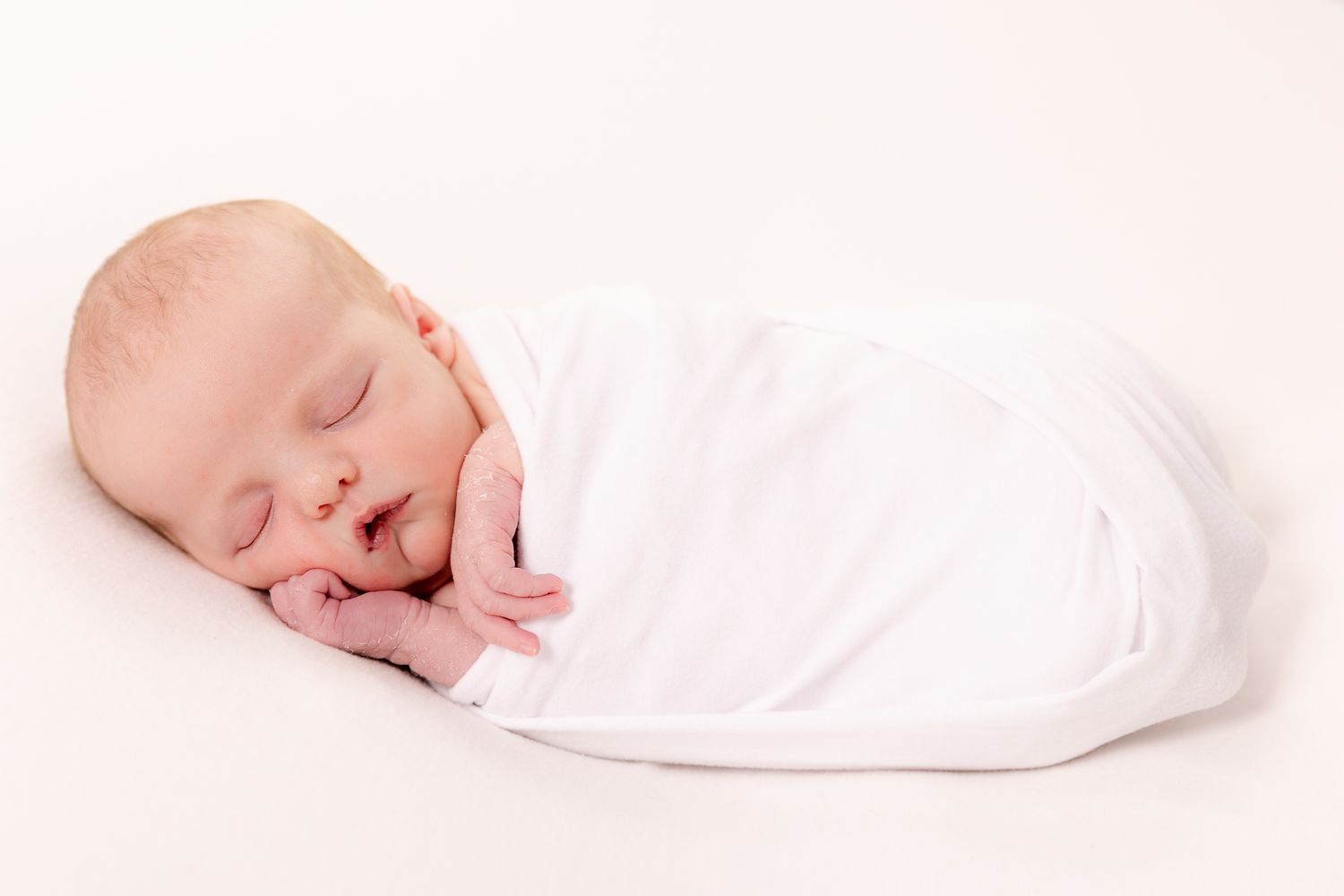 Newborn baby swaddled in a white wrap sleeping peacefully on a cream background in a studio portrait.