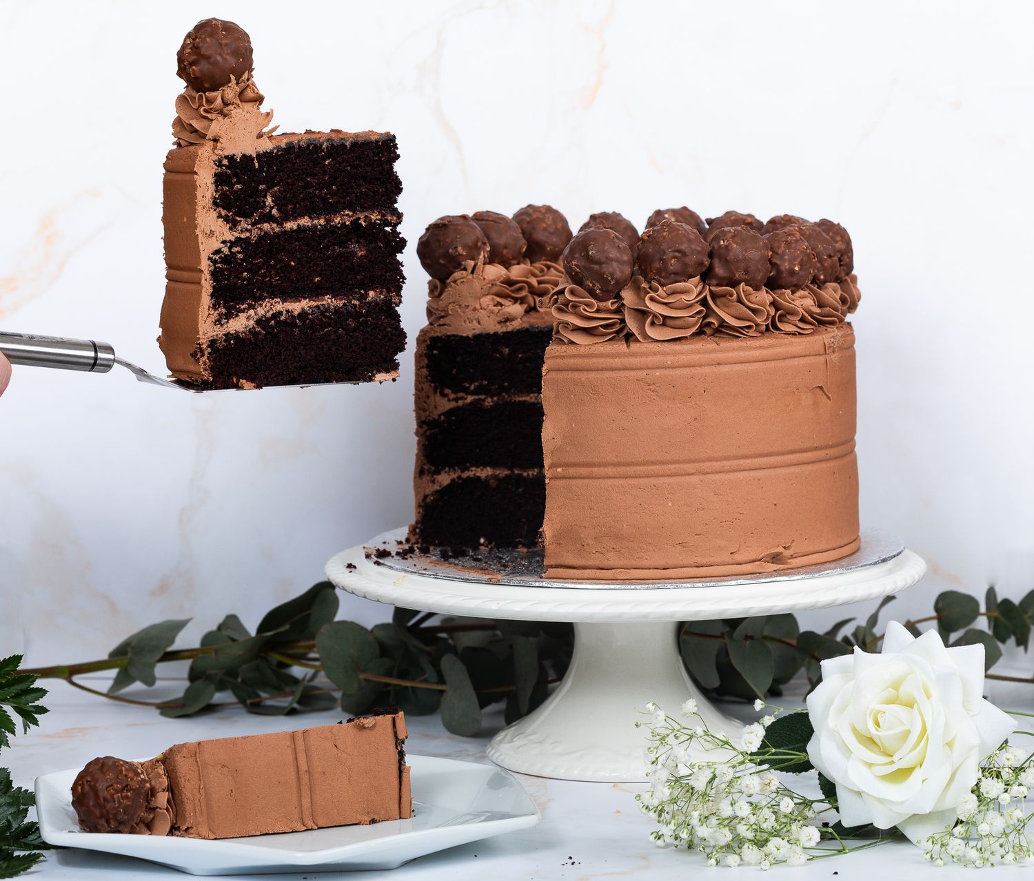 Rich chocolate layer cake being sliced on a white cake stand with white roses.