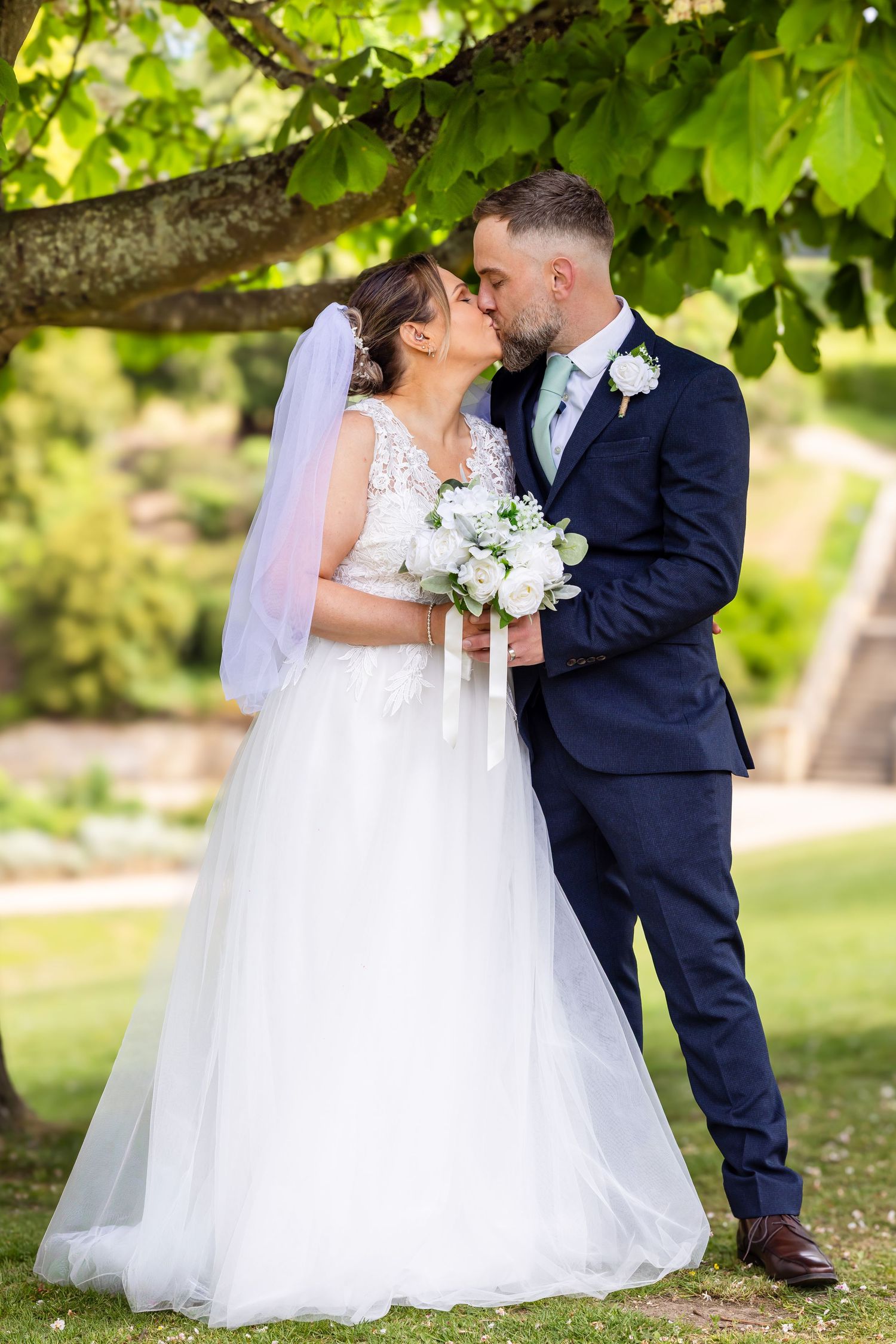 Bride and groom share a romantic kiss under a tree, bride holding white bouquet in a lace gown with veil.