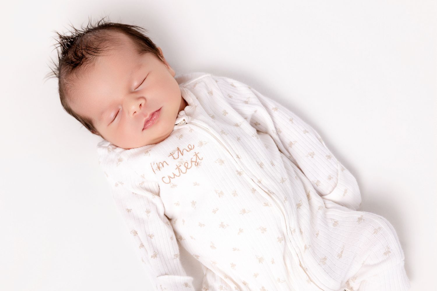 Sleeping newborn in white star print onesie with cute text, lying on white background.