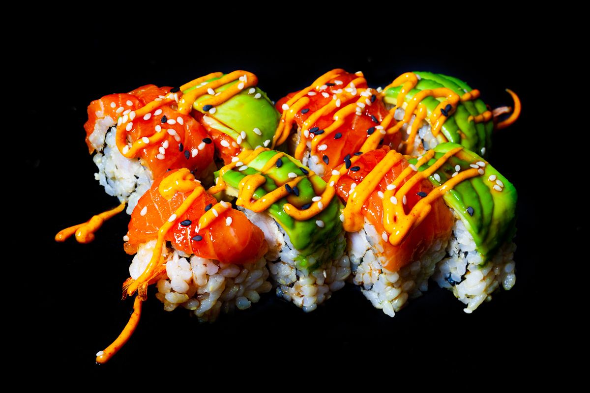 Colorful sushi rolls topped with avocado, sriracha, and sesame seeds on black background.