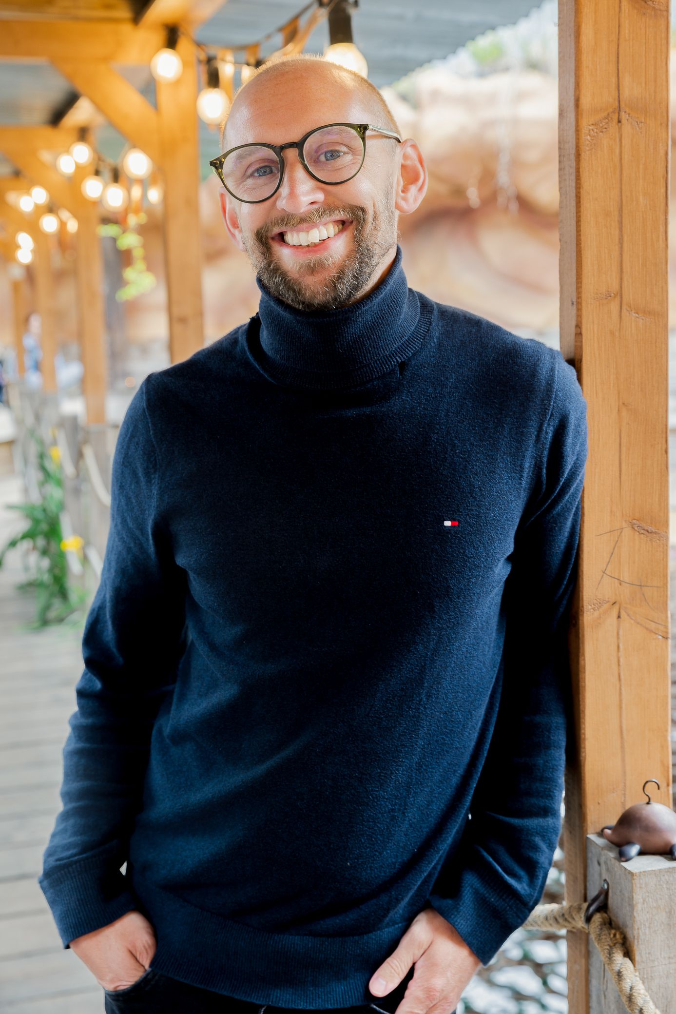 Bald smiling man with beard wearing glasses and navy turtleneck Tommy Hilfiger sweater leaning against wooden post.