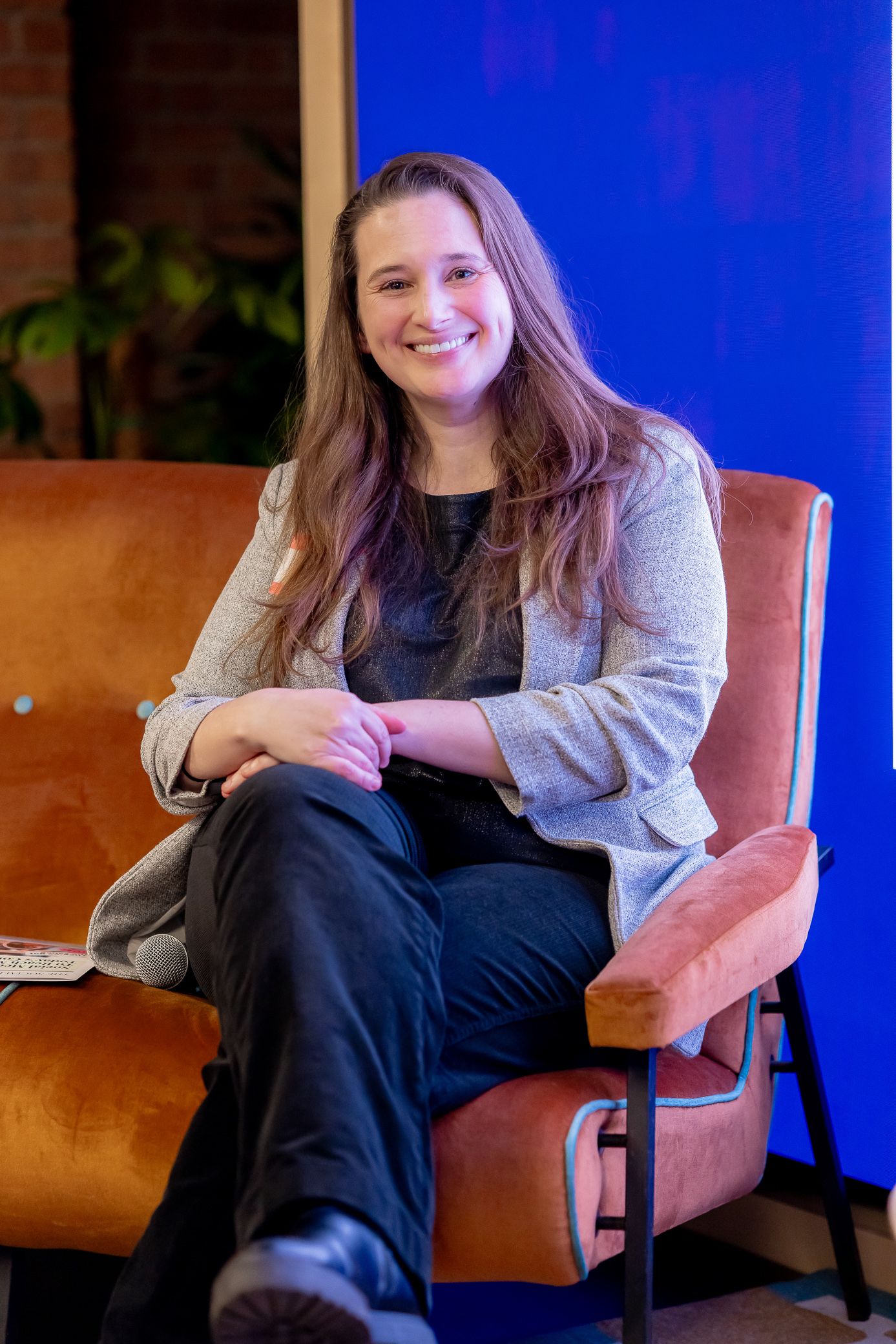 Woman with long hair smiling while seated in an orange chair at an indoor event.