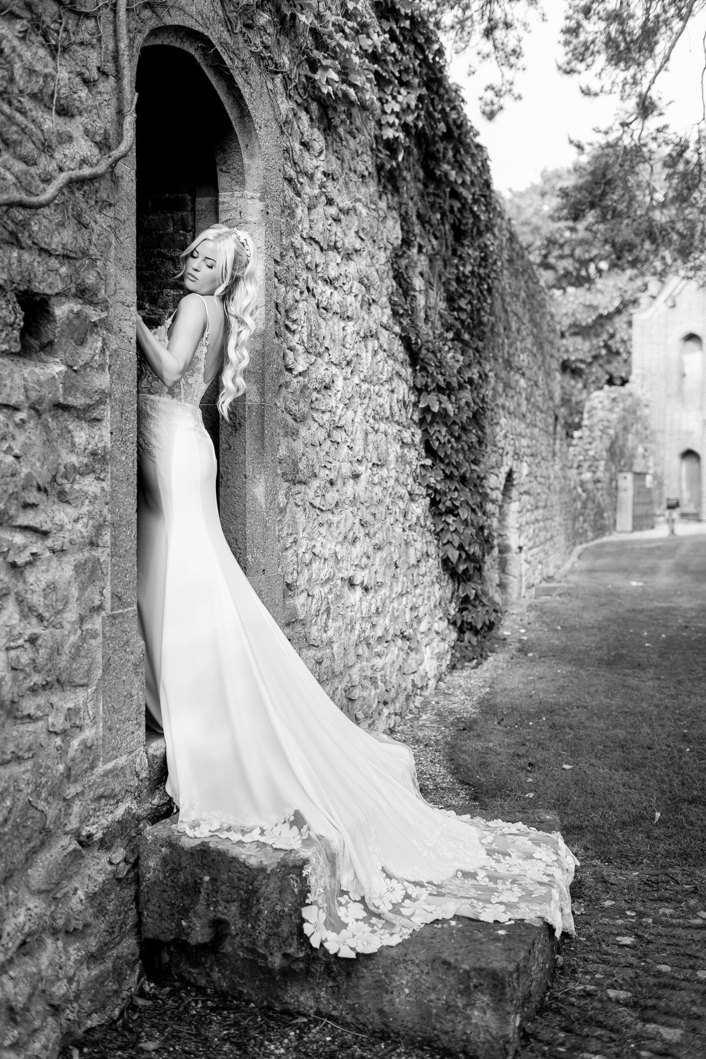 Black and white portrait of bride in lace gown with train at arched stone doorway.
