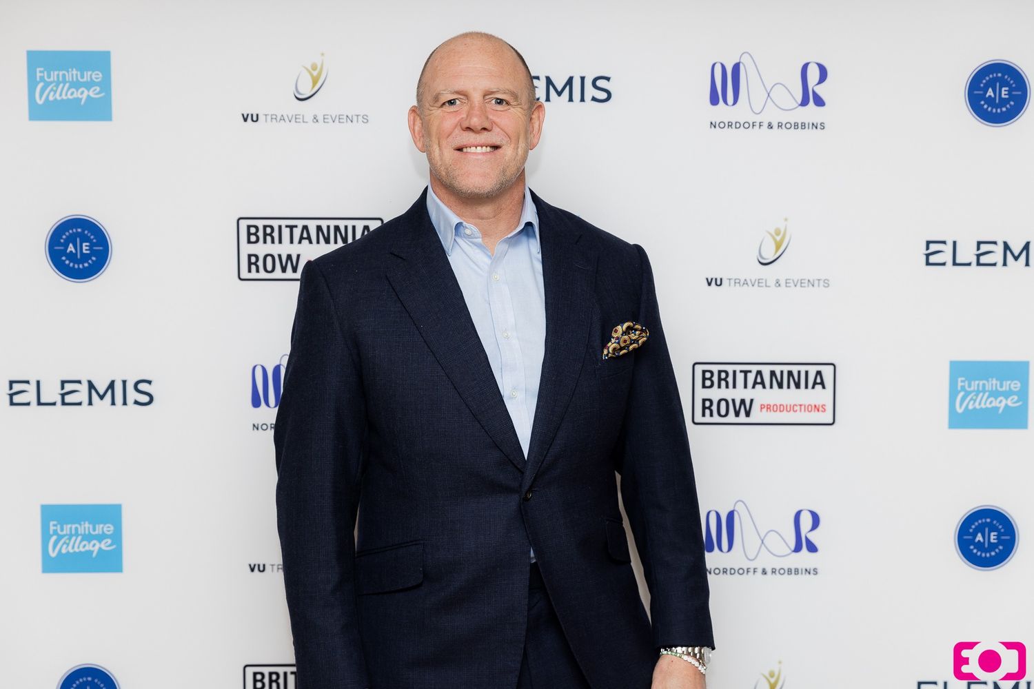 Man in navy blue suit with gold pocket square posing at Britannia Row Productions event with Elemis branding.
