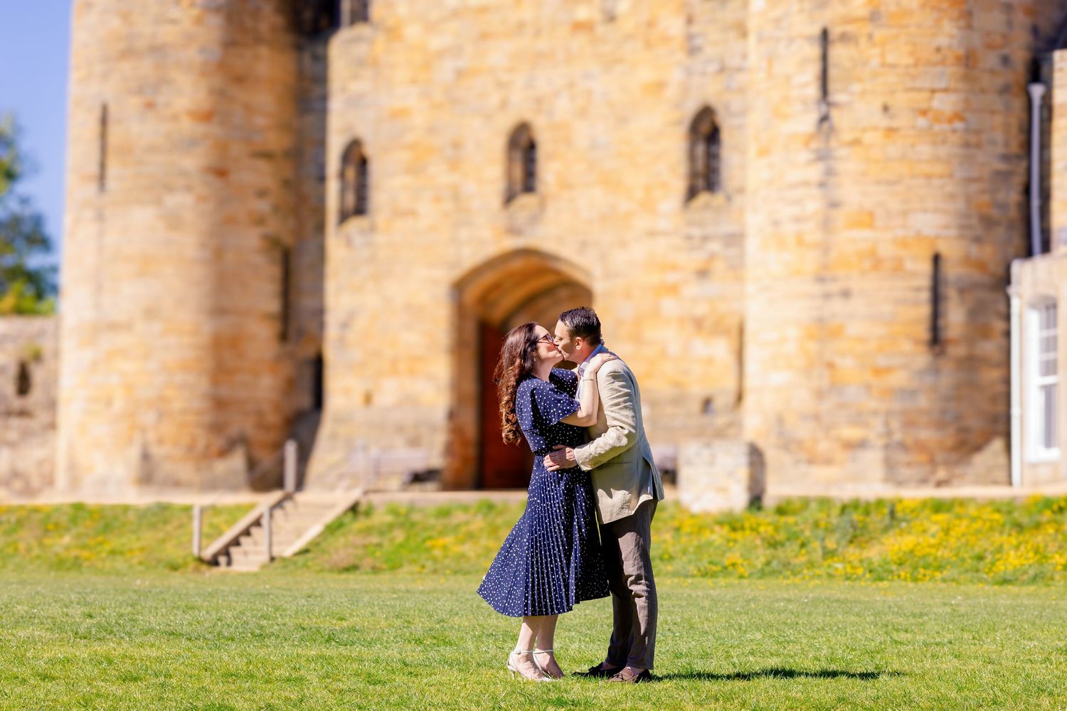 Couple sharing a romantic kiss on green lawn in front of a historic stone castle on a sunny day.
