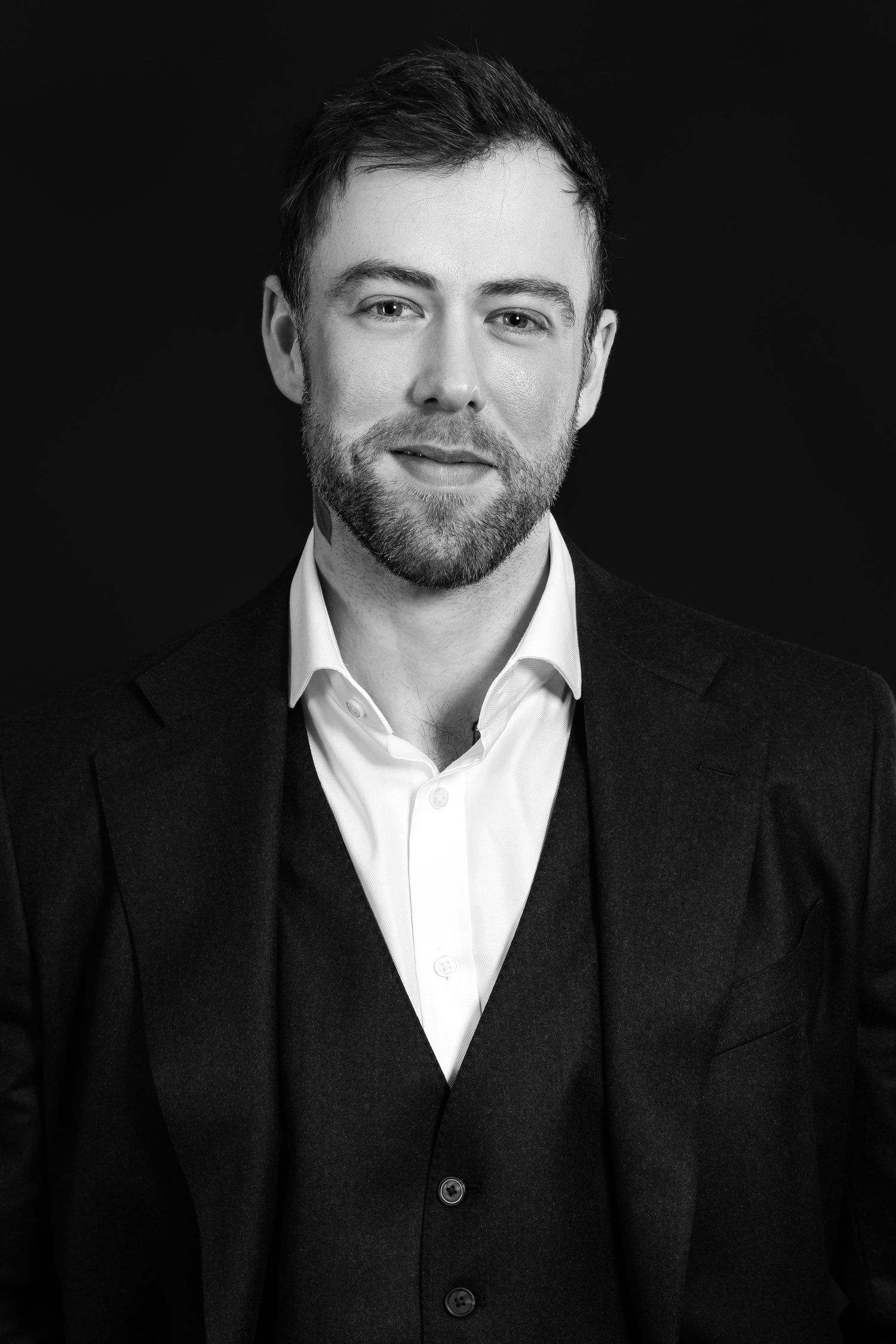 Black and white professional headshot of a bearded man in a suit and vest smiling confidently against dark background.
