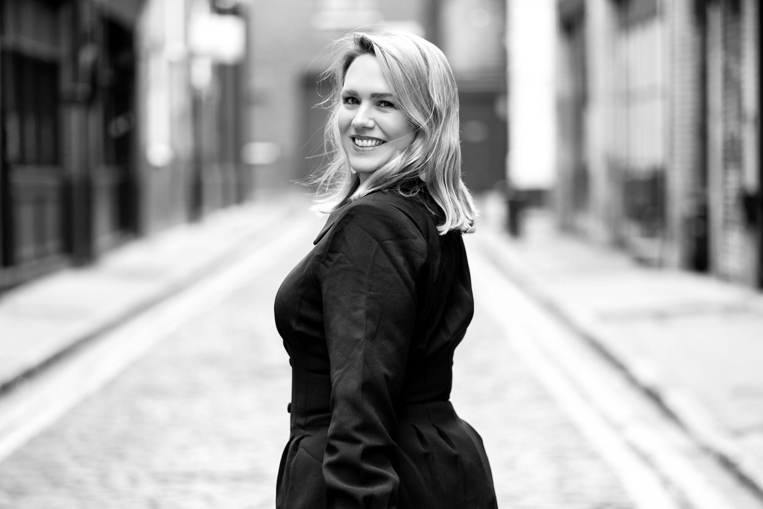 Black and white photo of a smiling blonde woman in a dark coat posing on a cobblestone alley street.
