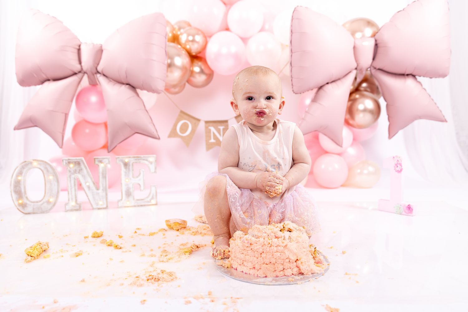 Baby girl celebrates first birthday smash cake session with pink balloons, bows, and ONE sign on white backdrop.