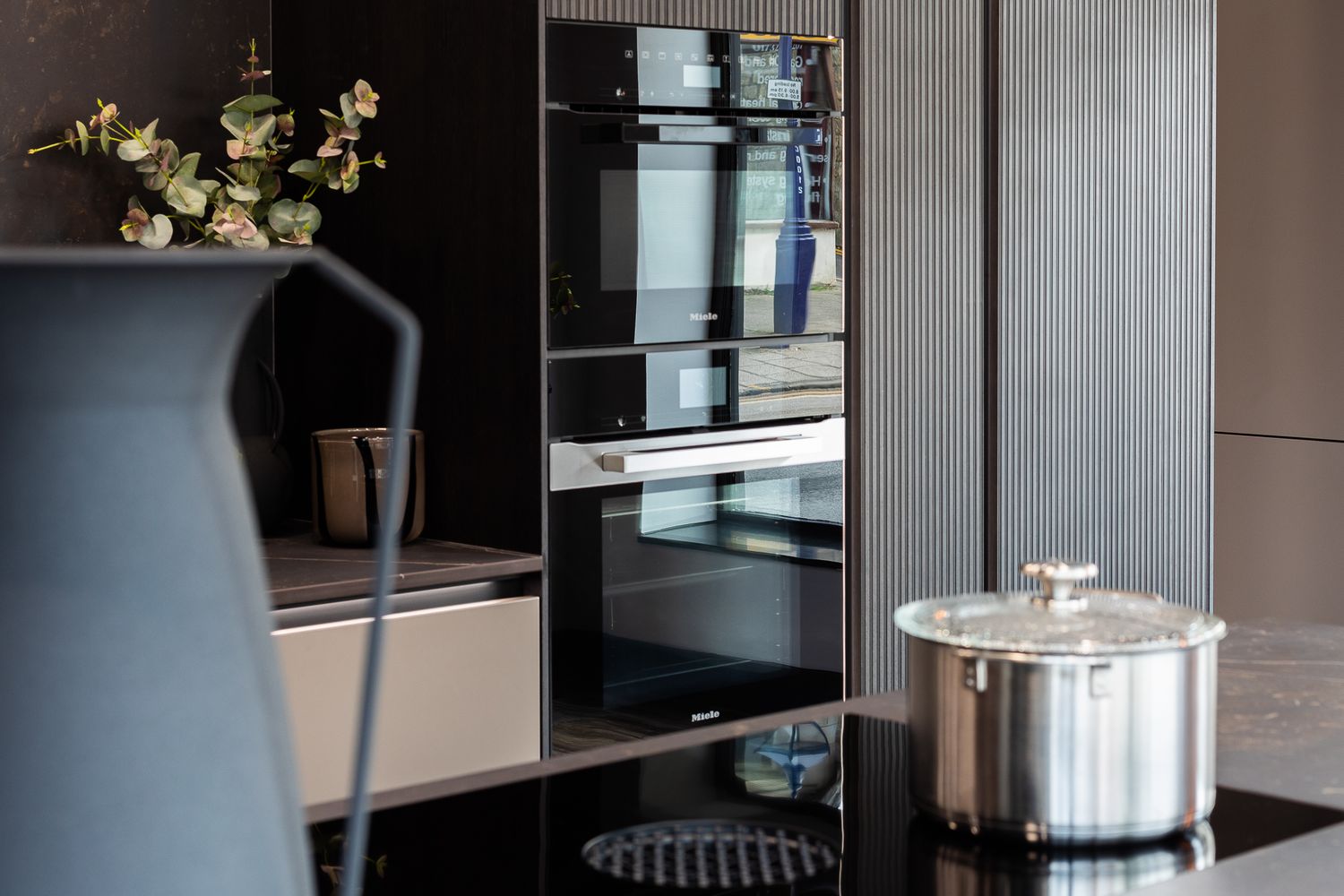 Modern black kitchen with built-in ovens, induction cooktop, and stainless steel pot on sleek dark cabinetry.