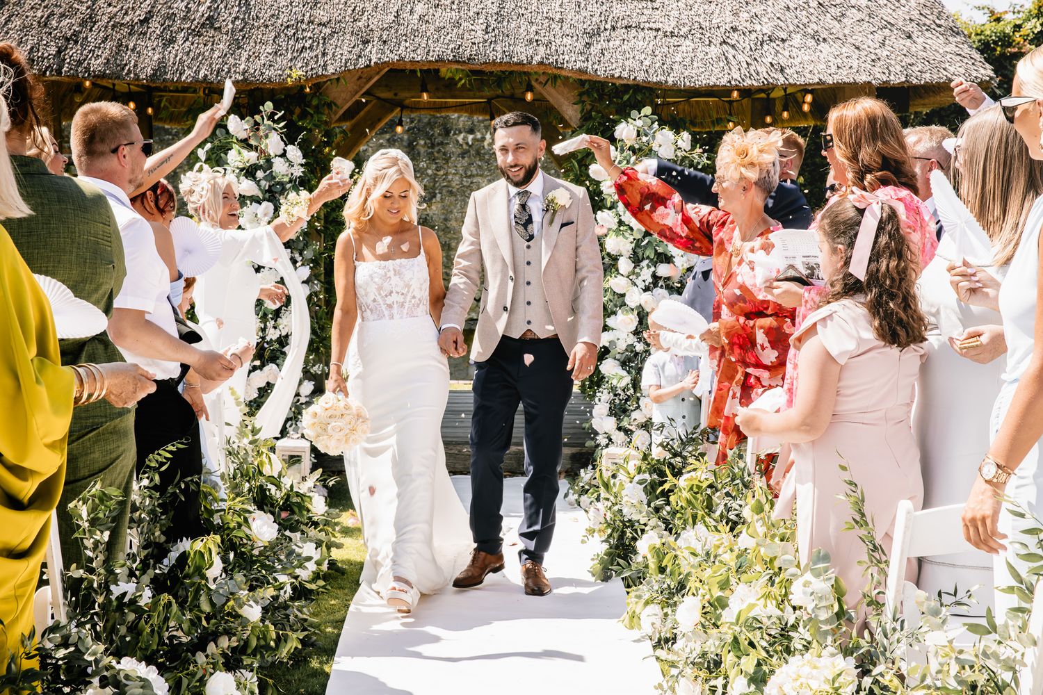 Bride and groom walking down floral aisle surrounded by cheering guests outdoors.