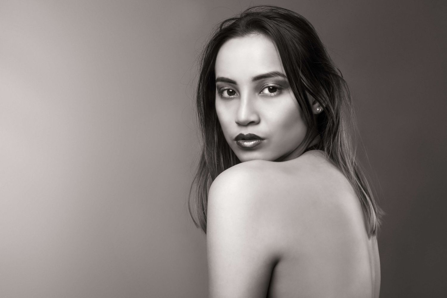Black and white artistic portrait of a young woman looking over her bare shoulder against a gray background.