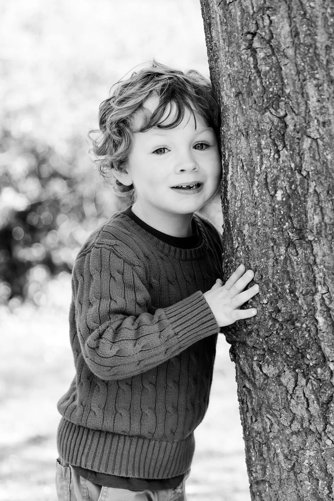 Black and white portrait of a smiling curly-haired boy leaning against a tree outdoors.