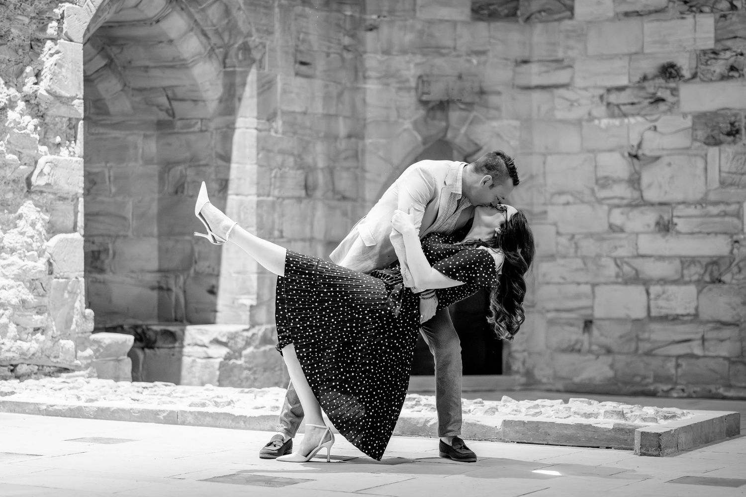Black and white photo of a couple sharing a romantic dip kiss in front of a rustic stone archway.
